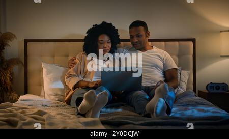 Happy family African American married couple relax at evening at home bedroom on bed man and woman at night browsing laptop using computer talking Stock Photo