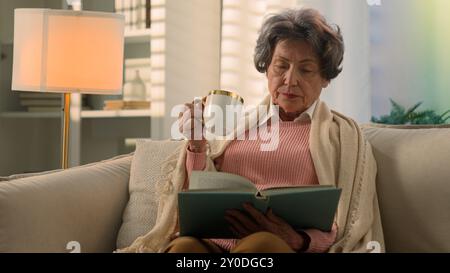 Calm serious Caucasian old woman covered with blanket on sofa drinking tea coffee with cup reading book senior mature lady elderly female retired Stock Photo