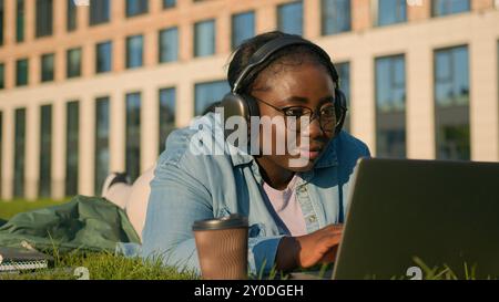 Happy student girl African American woman on lawn lying on green grass with wireless computer listening music in headphones study with laptop studying Stock Photo