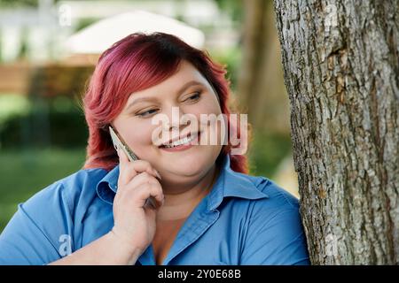 A plus size woman with bright pink hair speaks on her phone while standing by a tree. Stock Photo