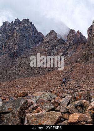 Traveler high in the mountains against the backdrop of majestic impregnable rocks. Climbing high in the mountains among rocks and stones. Trekking in Stock Photo