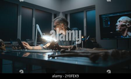 Archaeologist takes measurements of fossil bone under lamp, uses tablet computer. Scientist examines remains of ancient human in archaeological laboratory at night. Colleague works in the background. Stock Photo