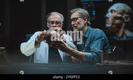 Two archaeologists take measurements of prehistoric human skull, conduct scientific research, use tablet computer. Scientists examine fossil remains in advanced archaeological laboratory at night. Stock Photo