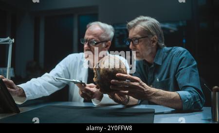 Two archaeologists measures skull of prehistoric extinct human, conduct historical and scientific research, use tablet computer. Scientists examine fossil remains in archaeological laboratory at night Stock Photo