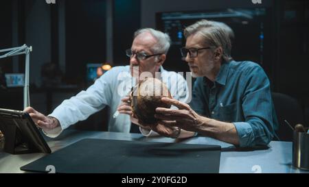 Two archaeologists talk and study skull of ancient extinct human, conduct historical and scientific research on fossil remains, use tablet computer. Scientists work in archaeological lab at night. Stock Photo