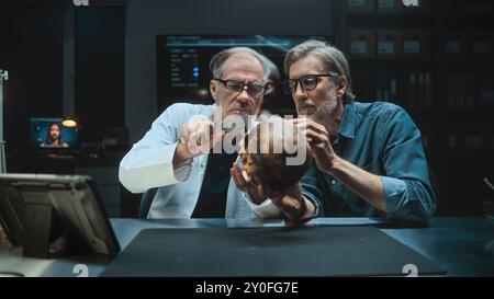 Two archaeologists talk and study skull of ancient extinct human, conduct historical and scientific research on fossil remains, use tablet computer. Scientists work in archaeological lab at night. Stock Photo