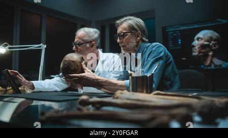 Two archaeologists measures skull of prehistoric extinct human, conduct historical and scientific research, use tablet computer. Scientists examine fossil remains in archaeological laboratory at night Stock Photo