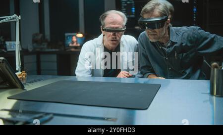 Two archaeologists wearing VR headsets conduct historical research of bones remains using augmented reality and tablet computer, have discussion. Scientists work in archaeological laboratory at night. Stock Photo