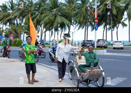 Nha Trang, Vietnam - April 22, 2024: Vietnamese elderly men pose on the street and smile. Rickshaw driver sits in his cart. Sea and palm trees are in Stock Photo