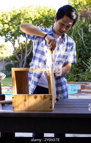 Using screwdriver, asian man assembling wooden box on outdoor table in backyard Stock Photo