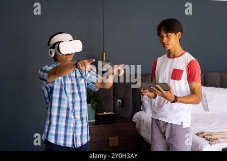Using VR headset, asian man interacting while another asian man holding tablet in bedroom Stock Photo