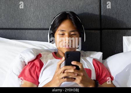 Listening to music with headphones, asian man using smartphone while relaxing in bed Stock Photo