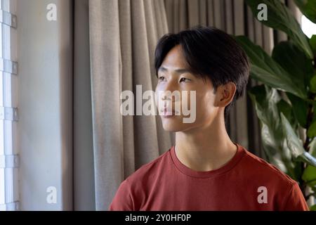 Young asian man in red shirt looking out window, contemplating in natural light, copy space Stock Photo