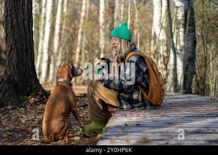 Female owner talks to dog sitting expectantly for treat on autumn forest walk. City escape with pet Stock Photo