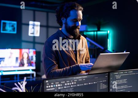 Smiling technician doing software debugging using laptop, checking glitches in lines of code displayed on PC monitors. Happy man walking in apartment fixing coding issues using notebook Stock Photo