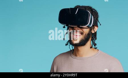 Middle Eastern man wearing virtual reality headset, doing swiping gestures. BIPOC person using high tech futuristic modern VR gadget, isolated over blue studio background, camera B Stock Photo