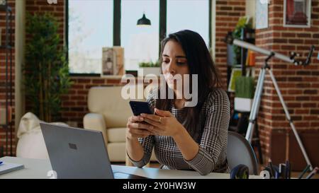 Smiling person checking mobile phone notifications at computer desk, taking break from work. Indian woman in cozy apartment pausing job tasks to read messages on cellphone, camera A zoom in Stock Photo
