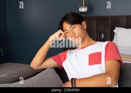 Pensive young asian man in casual wear sitting on couch, resting head on hand Stock Photo