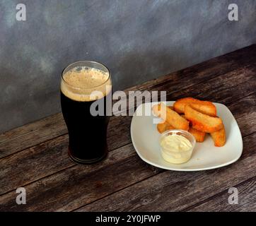 A plate of wheat croutons with sauce and a tall glass of dark beer with white foam on a dark wooden table. Close-up. Stock Photo