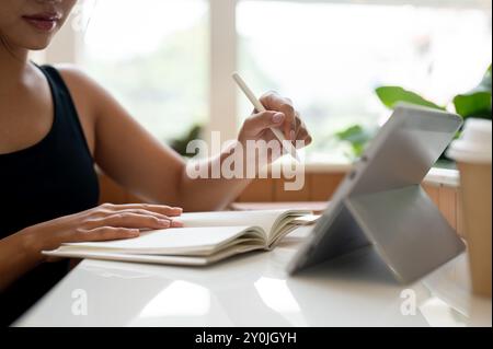 A cropped image of a beautiful Asian woman working remotely from a coffee shop, taking notes in a book while using her digital tablet. studying online Stock Photo