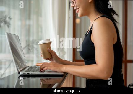 A cropped side view of an attractive Asian woman working remotely from a minimalist coffee shop, using a laptop while having coffee. people, lifestyle Stock Photo