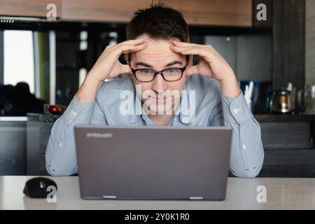 young man in eyeglasses holding laptop and looking extremely perplexed on home backdrop. Stock Photo