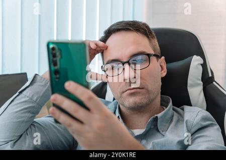 Portrait of a worried young man sitting in the office at the table, holding his head with his hand, looking sadly at the phone. Bored, received bad ne Stock Photo