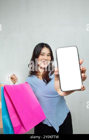 woman standing smiling with mobile phone, blue stylish casual Asian girl showing mobile phone blank screen, white background studio Stock Photo
