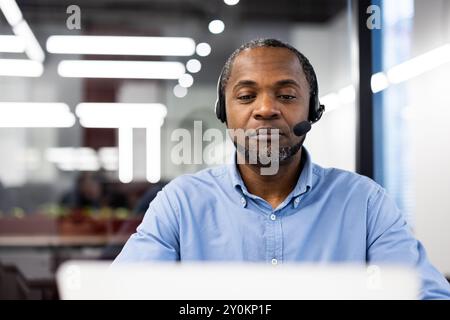Serious businessman using headset for virtual meeting. Professional appearance, dressed in blue shirt, concentrated on work. Sitting in modern office environment communicating online Stock Photo