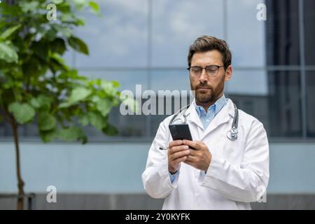 Male doctor standing outdoors wearing white coat and stethoscope, using phone. Medical professional looking serious, focused on digital device in urban setting. Concept of healthcare, technology Stock Photo