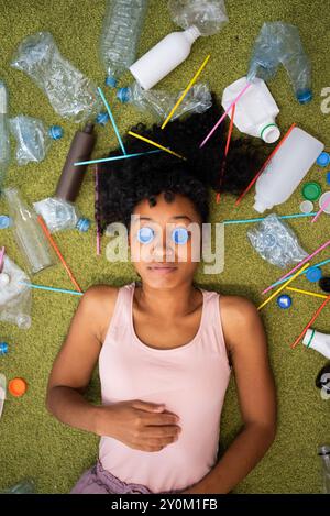 African-American woman is lying on green carpet, and plastic waste is scattered around, eyes are covered with bottle caps. Impact of plastic pollution on environment Stock Photo