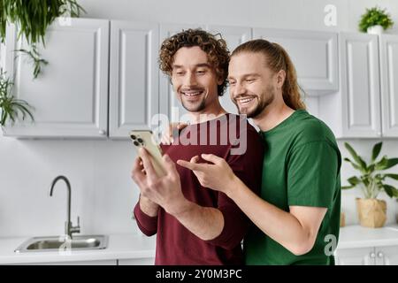Two men joyfully engage with their smartphone in a modern kitchen. Stock Photo