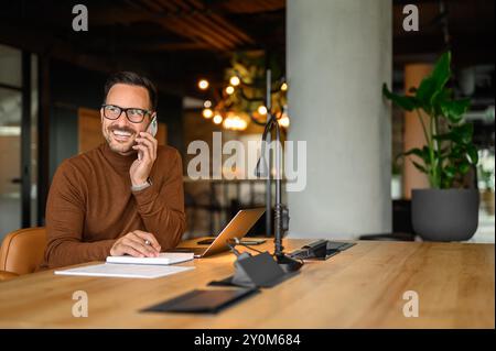 Male programmer smiling and discussing project over phone call while working on laptop at desk in office Stock Photo