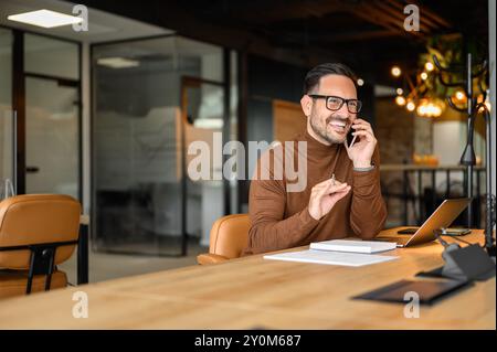 Cheerful male professional with laptop and notepad on desk talking over smart phone at his workplace Stock Photo