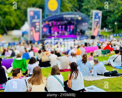 Large crowd gathered in park for outdoor music festival. Colorful blankets and chairs spread across grass while people enjoy live performance in sunlight. Stock Photo