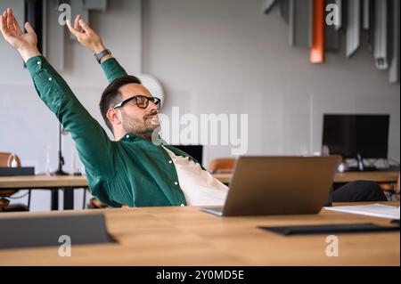 Exhausted male manager with eyes closed stretching hands and relaxing at his desk in modern office Stock Photo
