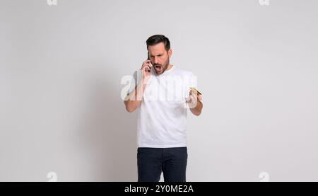 Shocked angry businessman with credit card talking on smart phone while standing on white background Stock Photo