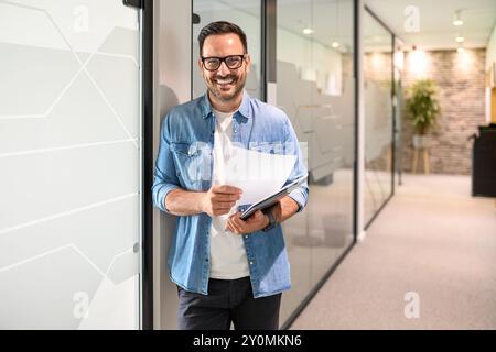 Portrait of confident male programmer with laptop and document standing by glass wall in modern office Stock Photo