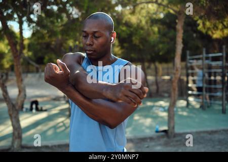 African athletic man dressed in sleeveless blue shirt, black shorts do shoulder stretching warming up exercises in park, sunny summer day. Sport, heal Stock Photo