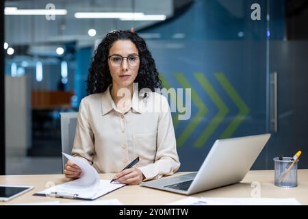 Hispanic businesswoman in office setting examining documents and using laptop. Professional and focused, showcasing concept of business and modern work environment. Stock Photo