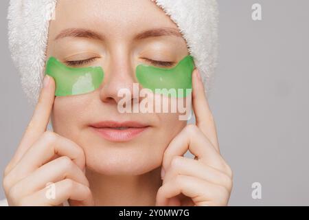 A woman with a towel on her head and a robe smiles as she applies green gel patches under her eyes facial cosmetology concept Stock Photo