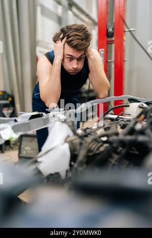 Worried mechanic having a stressful day at car repair shop, holding head in hands while looking at car engine. Confused repairman unsure of how to fix Stock Photo