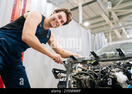 Low-angle view of cheerful mechanic male in blue overalls happily using tools to repair engine of car at auto repair shop, smiling looking at camera. Stock Photo