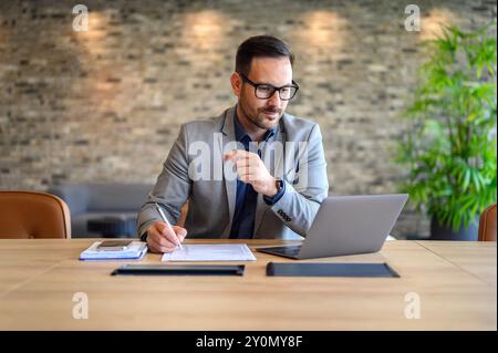 Focused male project manager writing on paper while doing online research over laptop at desk in office Stock Photo