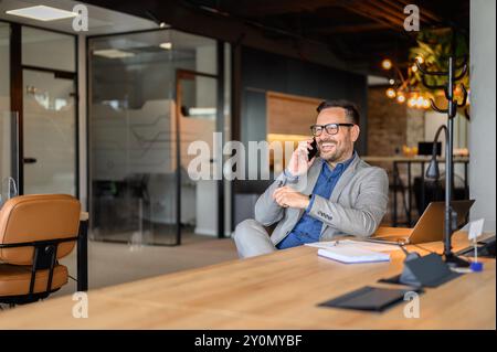 Cheerful male professional with laptop and notepad on desk talking over smart phone at his workplace Stock Photo