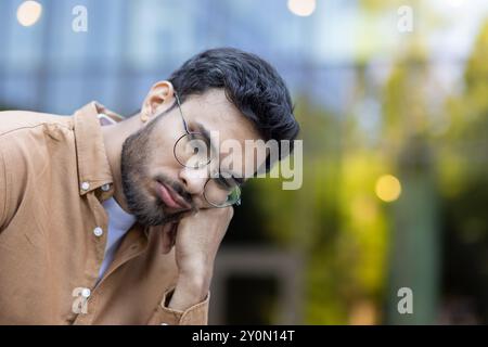 Thoughtful young man wearing glasses in casual shirt leans on hand, appearing deep in reflection. Background suggests office environment, depicting moment of contemplation during break from work. Stock Photo
