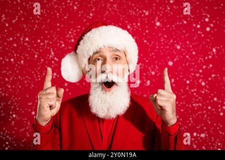 Photo of shocked funky santa claus indicate forefingers up empty space open mouth wear x-mas hat suit on red color background Stock Photo
