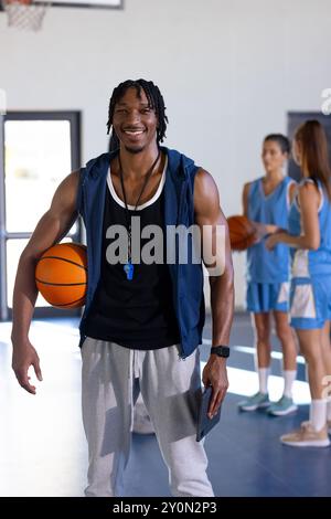 male african American basketball coach holding ball and whistle, smiling in gym with female players Stock Photo