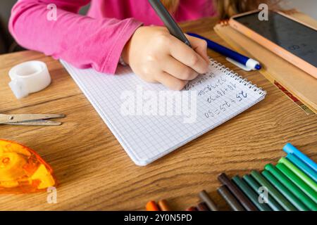Child doing math exercises in notebook using smartphone with stationery on wooden table. Girl hand writing mathematical equations as school homework Stock Photo
