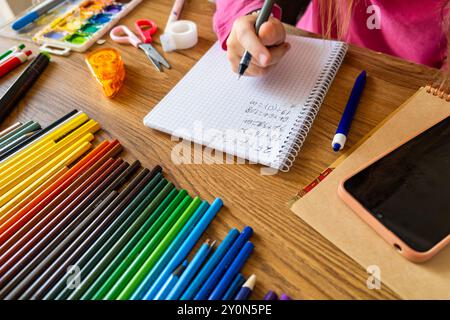 Child doing math test in notebook using smartphone with stationery on wooden table. Girl hand writing mathematical exercises as school homework Stock Photo